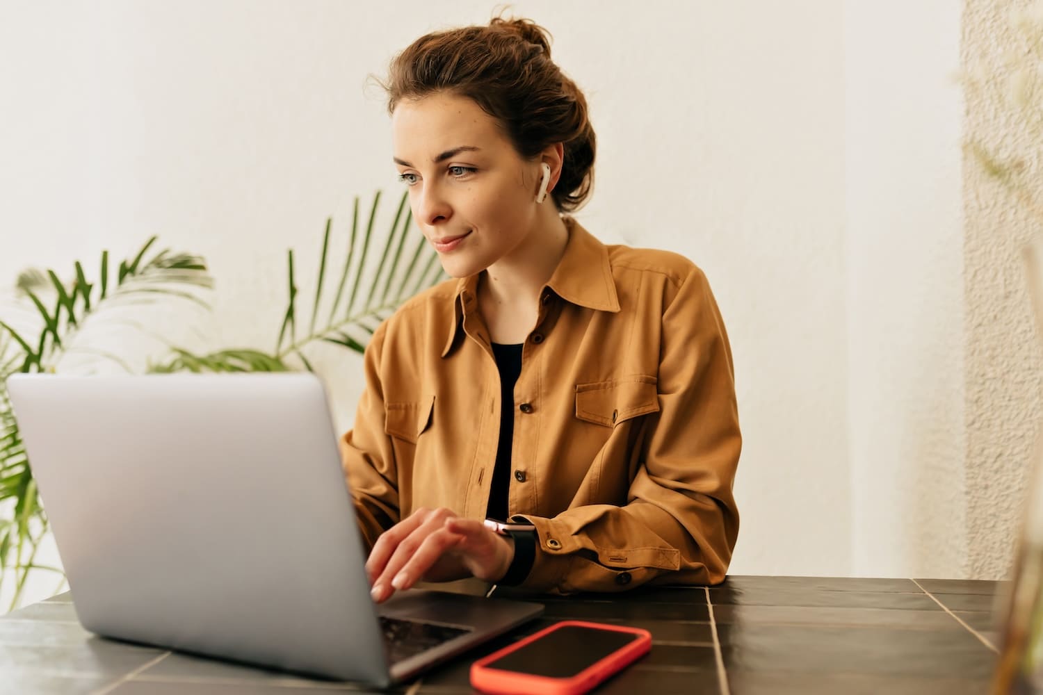 Nebenbeschäftigung_HR Nebenbeschäftigung: Frau arbeitet am Laptop, trägt beiges Hemd, grüner Hintergrund mit Pflanzen, moderne und ruhige Atmosphäre.